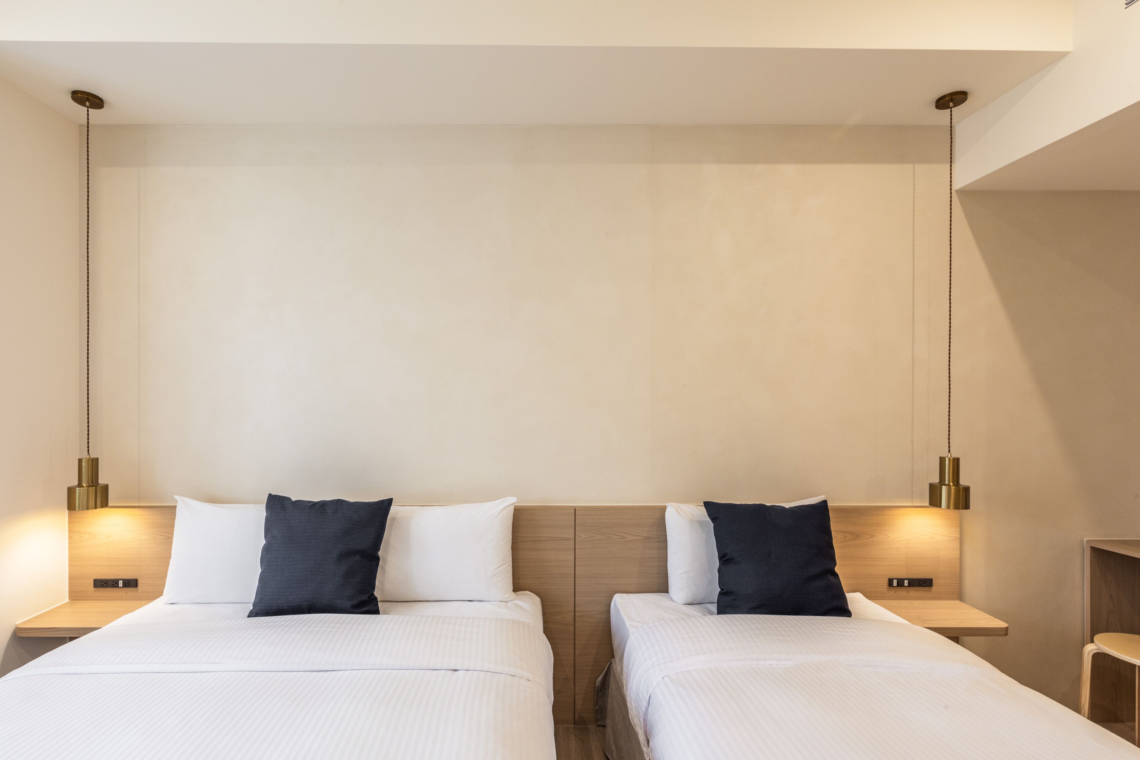 A hotel room with two beds, each with a wooden headboard, white bedding, and a dark pillow. There are bedside tables with lamps and a small wooden chair.