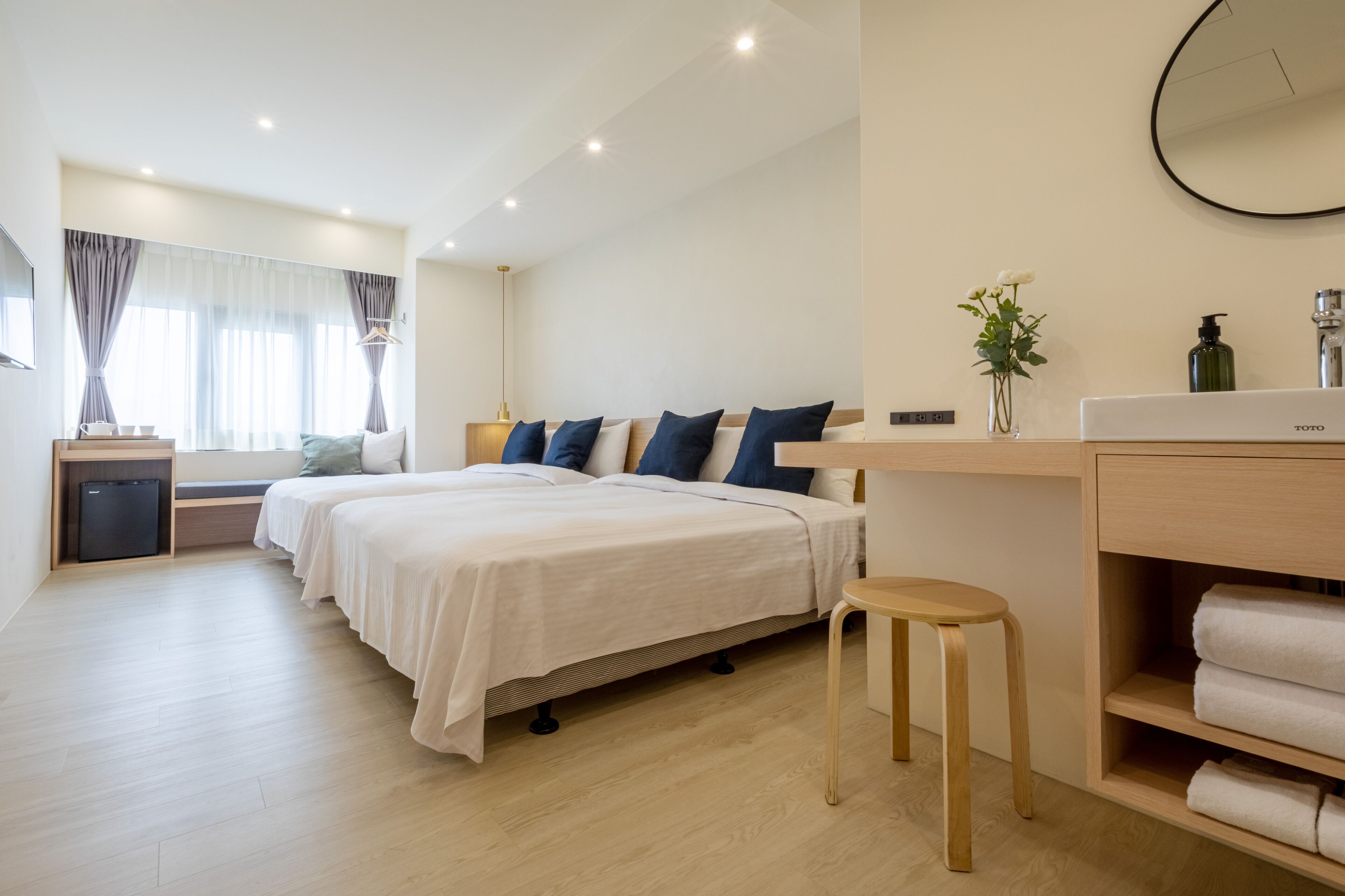 A modern hotel room with two beds, a small desk, a vanity with a mirror, and a wooden stool.