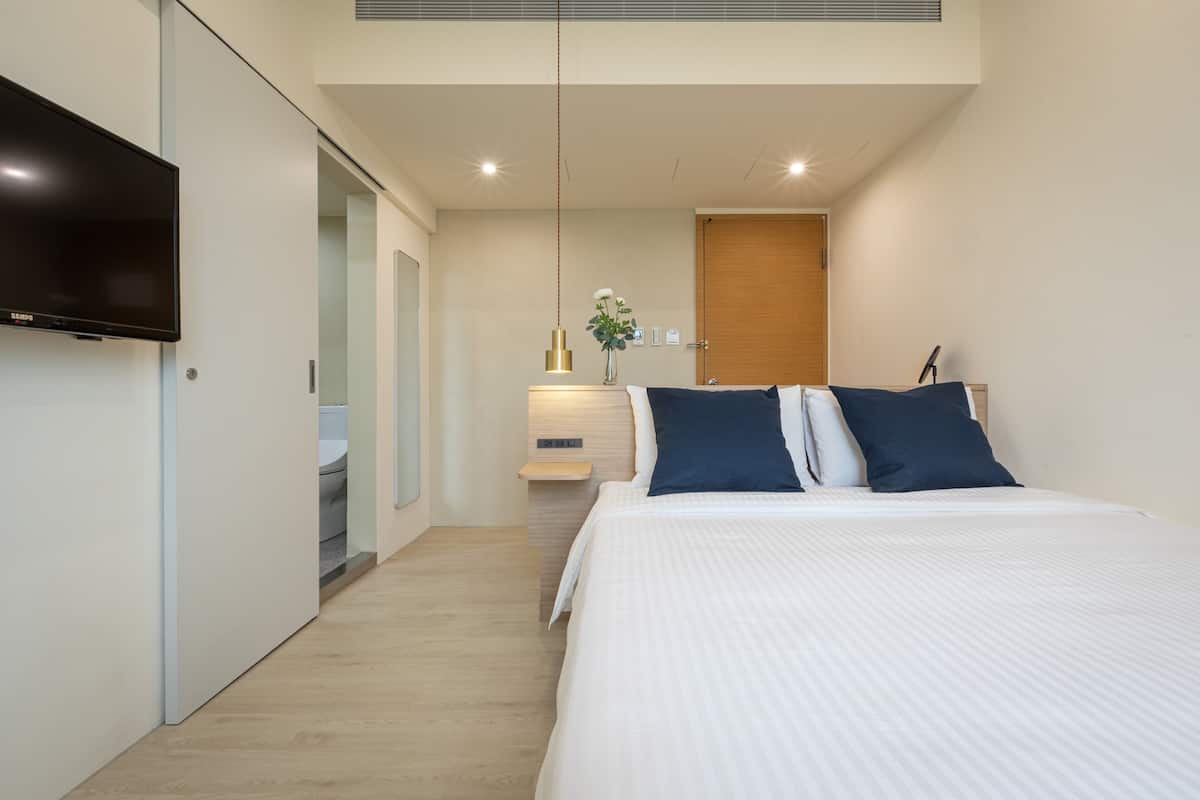 A modern hotel room with a large bed, a flat-screen TV mounted on the wall, a bedside table with a lamp, and a wooden door.