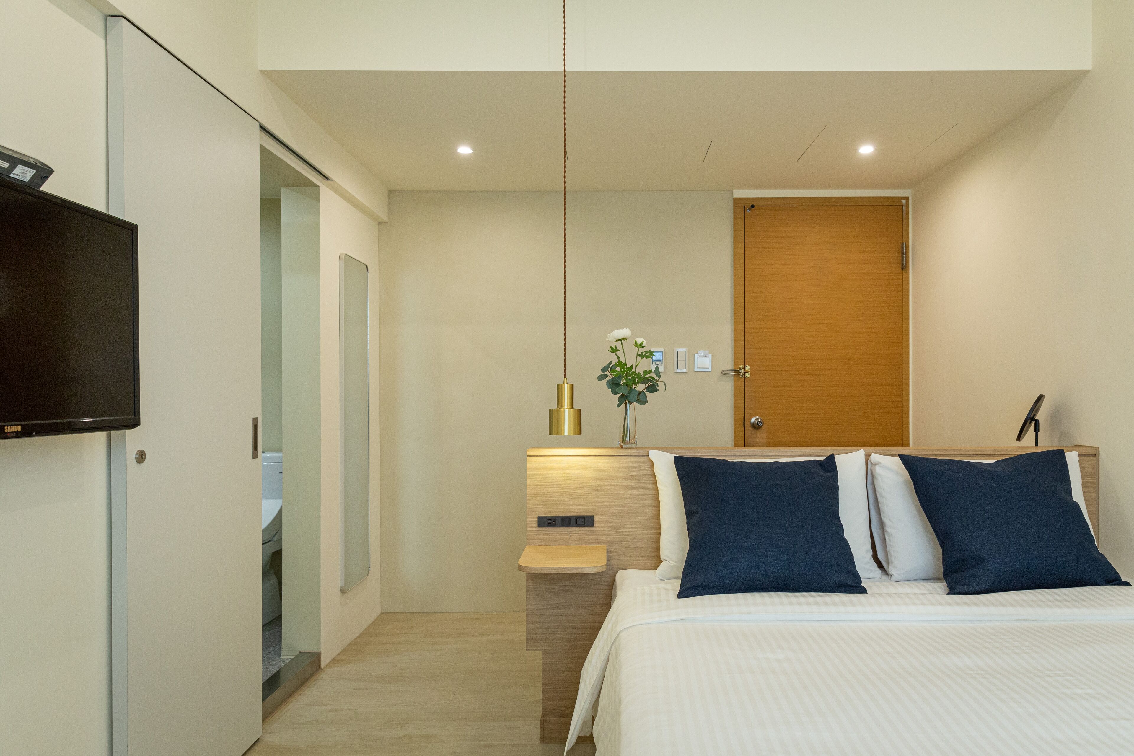 A modern hotel room with a bed, a TV, a wooden door, and a small table with a vase.