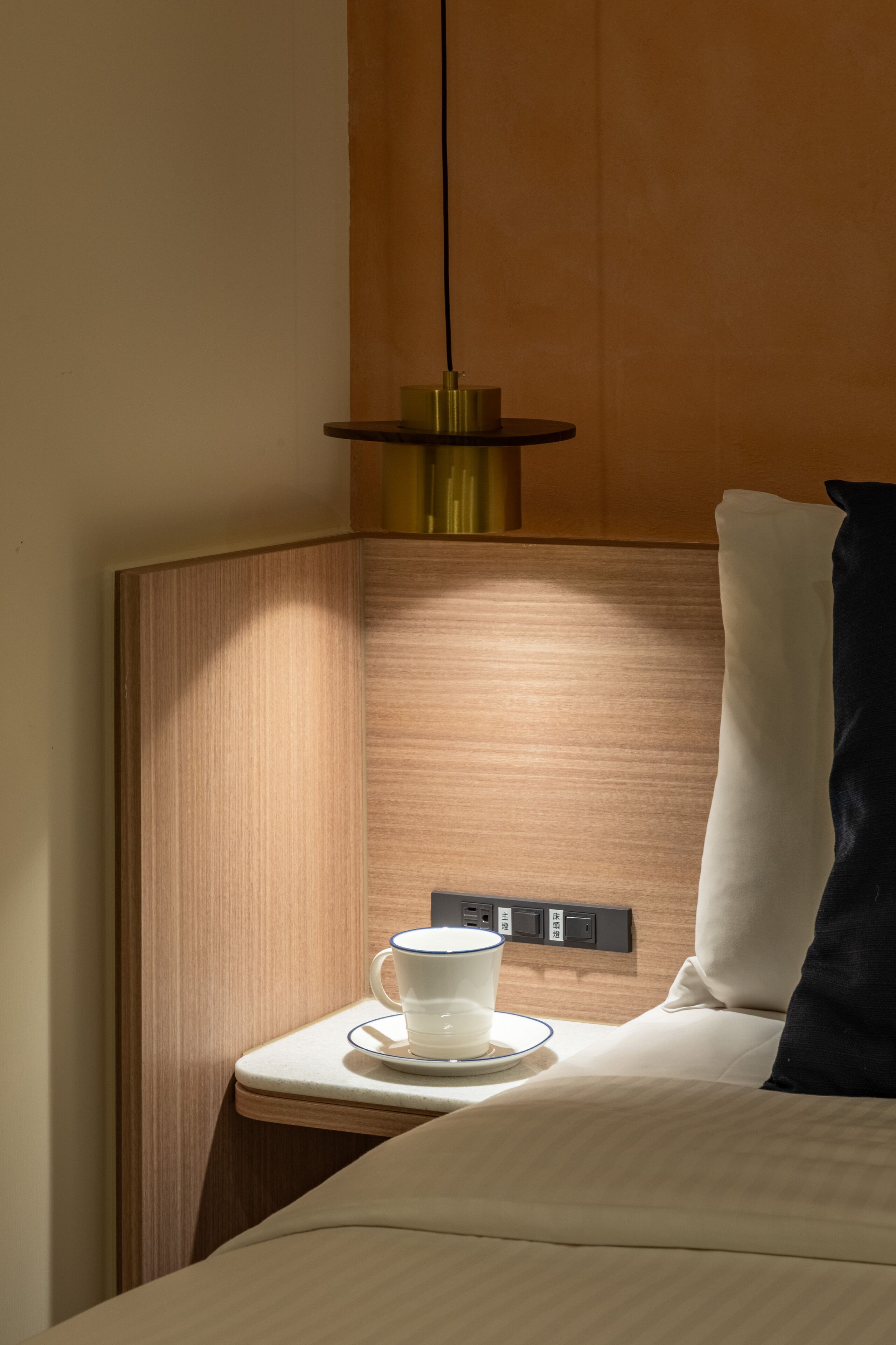 Eine Ecke eines Hotelzimmers mit einem hölzernen Kopfteil, einem Nachttisch mit Tasse und Untertasse sowie einer Steckdose.
