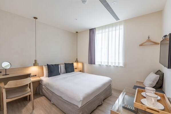 A modern hotel room with a large bed, bedside tables, a desk, a chair, a TV, and a window with curtains.