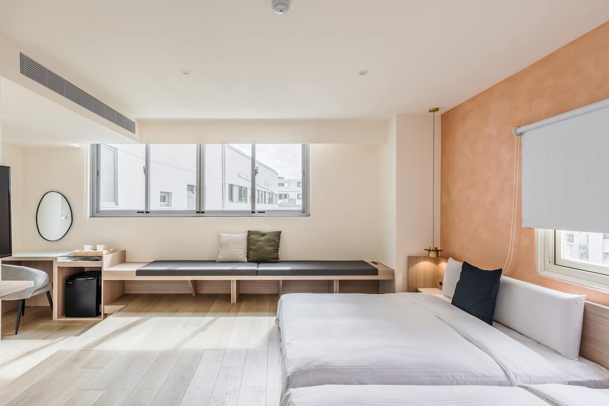 A modern hotel room with a large bed, a desk with a chair, a bench with cushions, a small refrigerator, and a large window offering a view of the city.