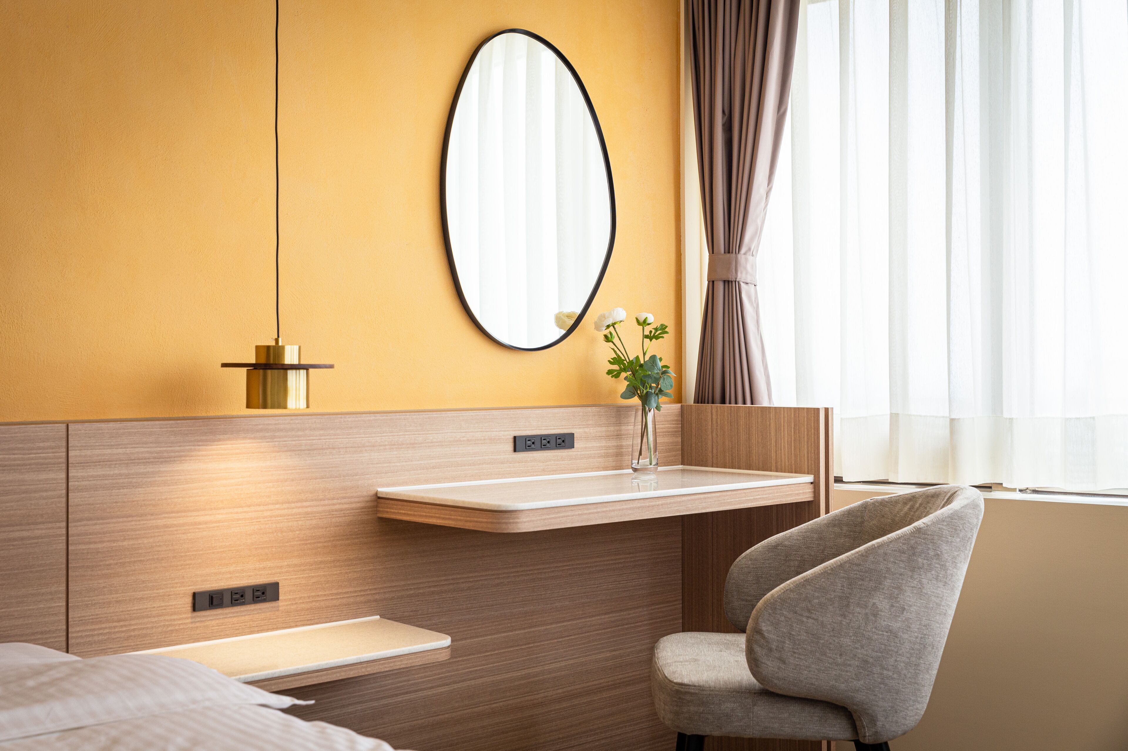 A modern hotel room with a wooden headboard, a bedside table, a chair, a mirror, and a window with curtains.