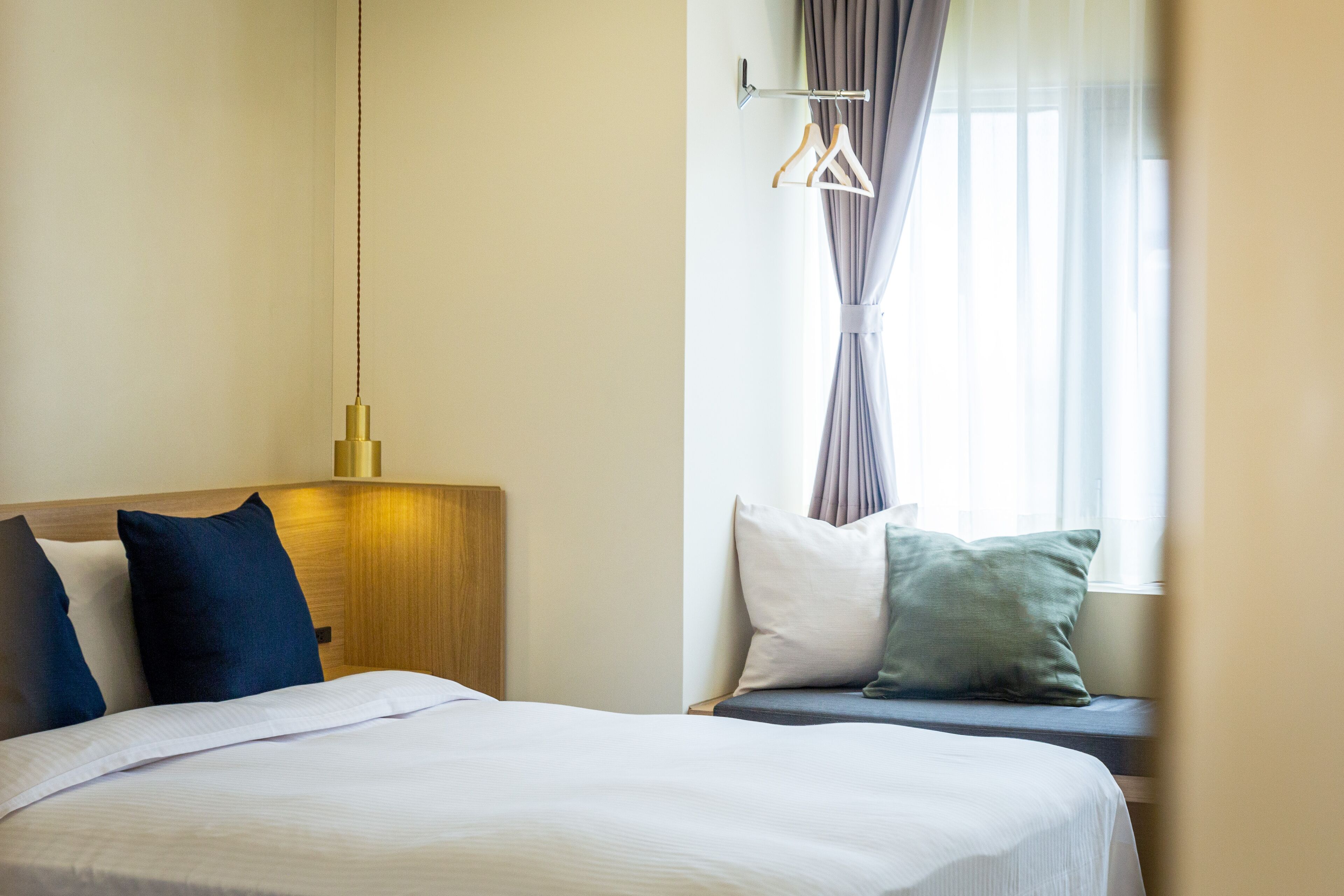 Een hotelkamer met een bed, een hanglamp, een raam met gordijnen en een kapstok aan de muur.