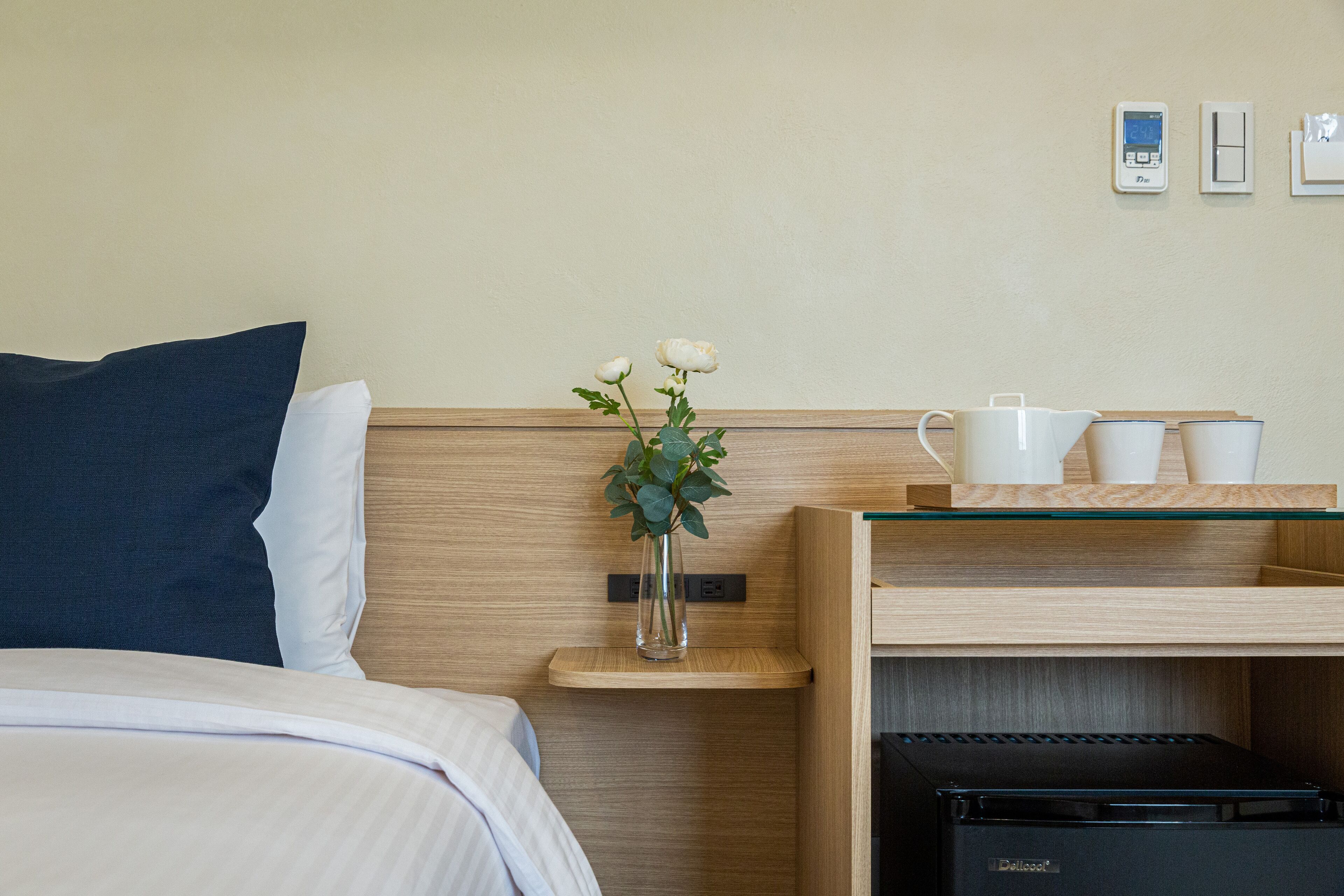 A neatly made hotel room with a bed, a bedside table with a vase of flowers, a coffee maker, and a small shelf with cups.