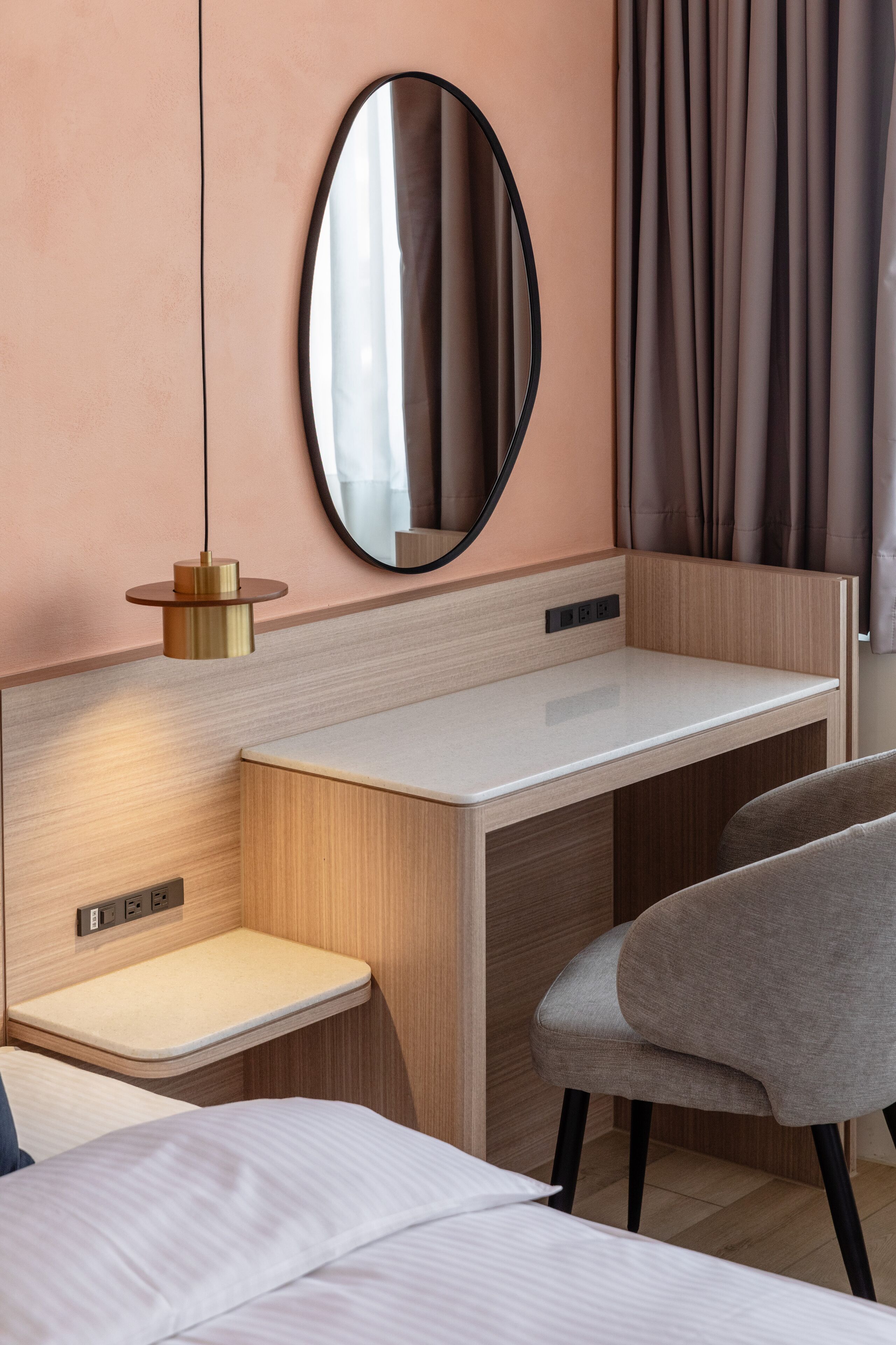 A modern hotel room with a wooden desk, a chair, a bedside table, and a large mirror.