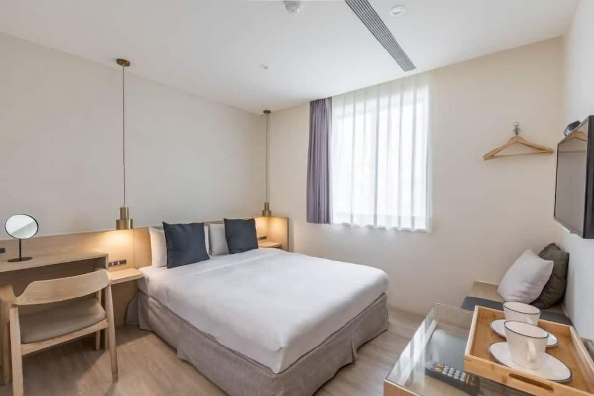 A modern hotel room with a large bed, bedside table, lamp, chair, and a small desk with a mirror.