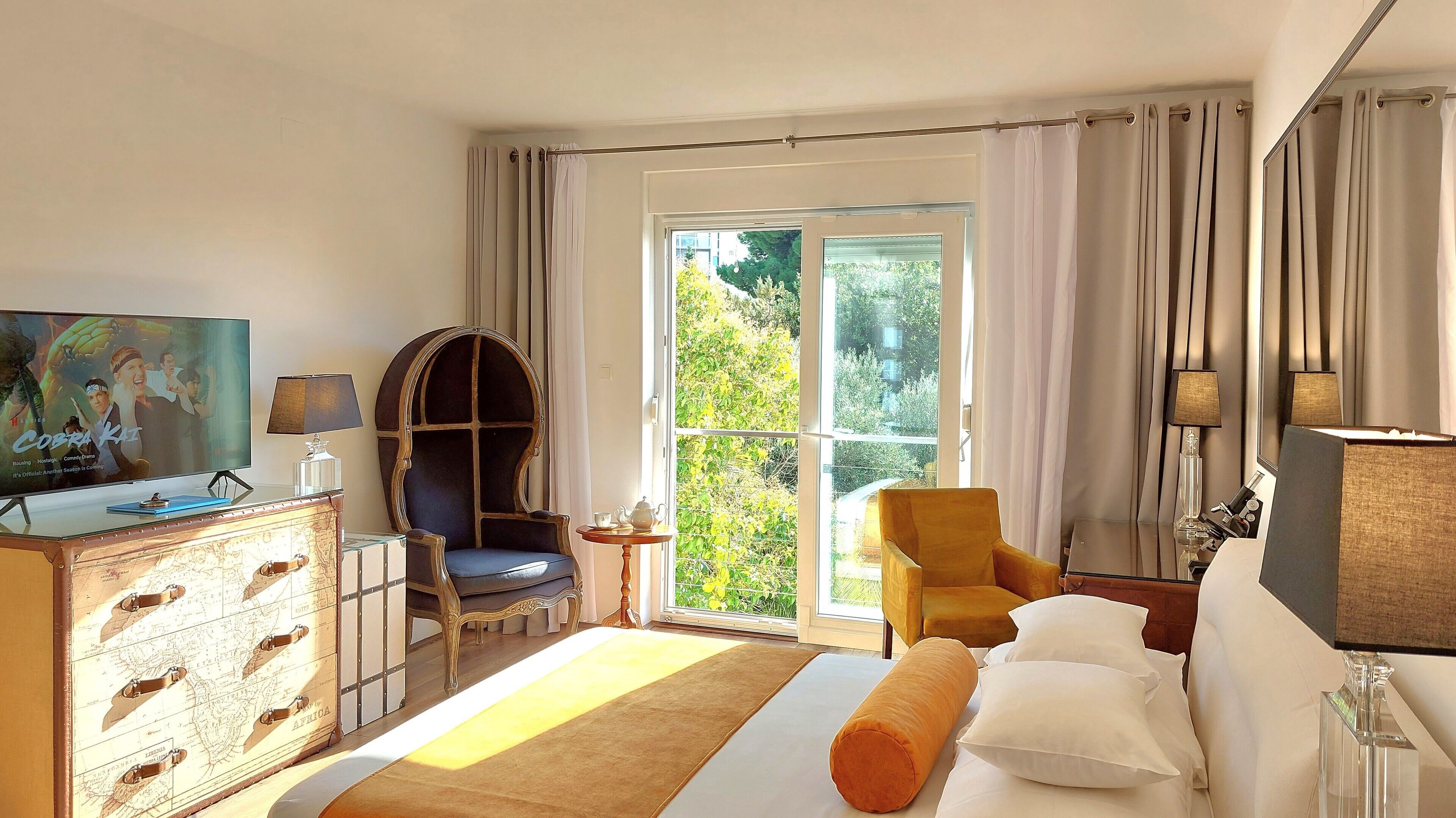 Habitación de hotel con una cama grande, televisor, escritorio con lámpara, dos sillas, mapa en la cómoda y puerta corredera de vidrio al exterior.