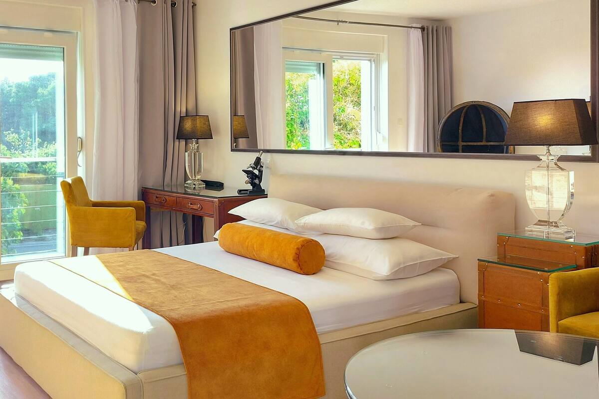 Habitación de hotel con cama, escritorio, silla, lámpara y vista a la vegetación.