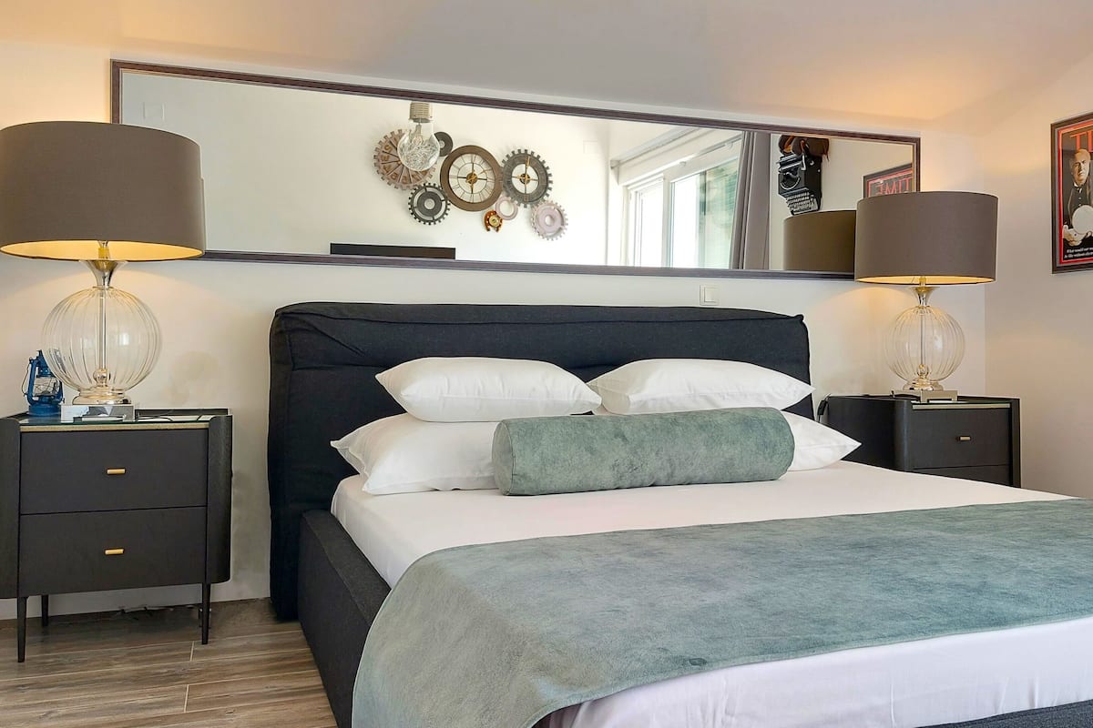 Una cama bien hecha con un plaid verde, dos mesitas de noche con lámparas, un espejo grande sobre la cama y una revista TIME en la pared.