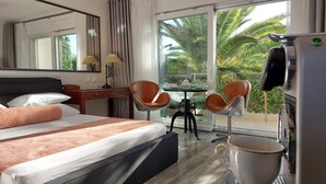 Habitación de hotel con cama, dos sillas, una mesita con tetera y vistas a palmeras a través de la ventana.
