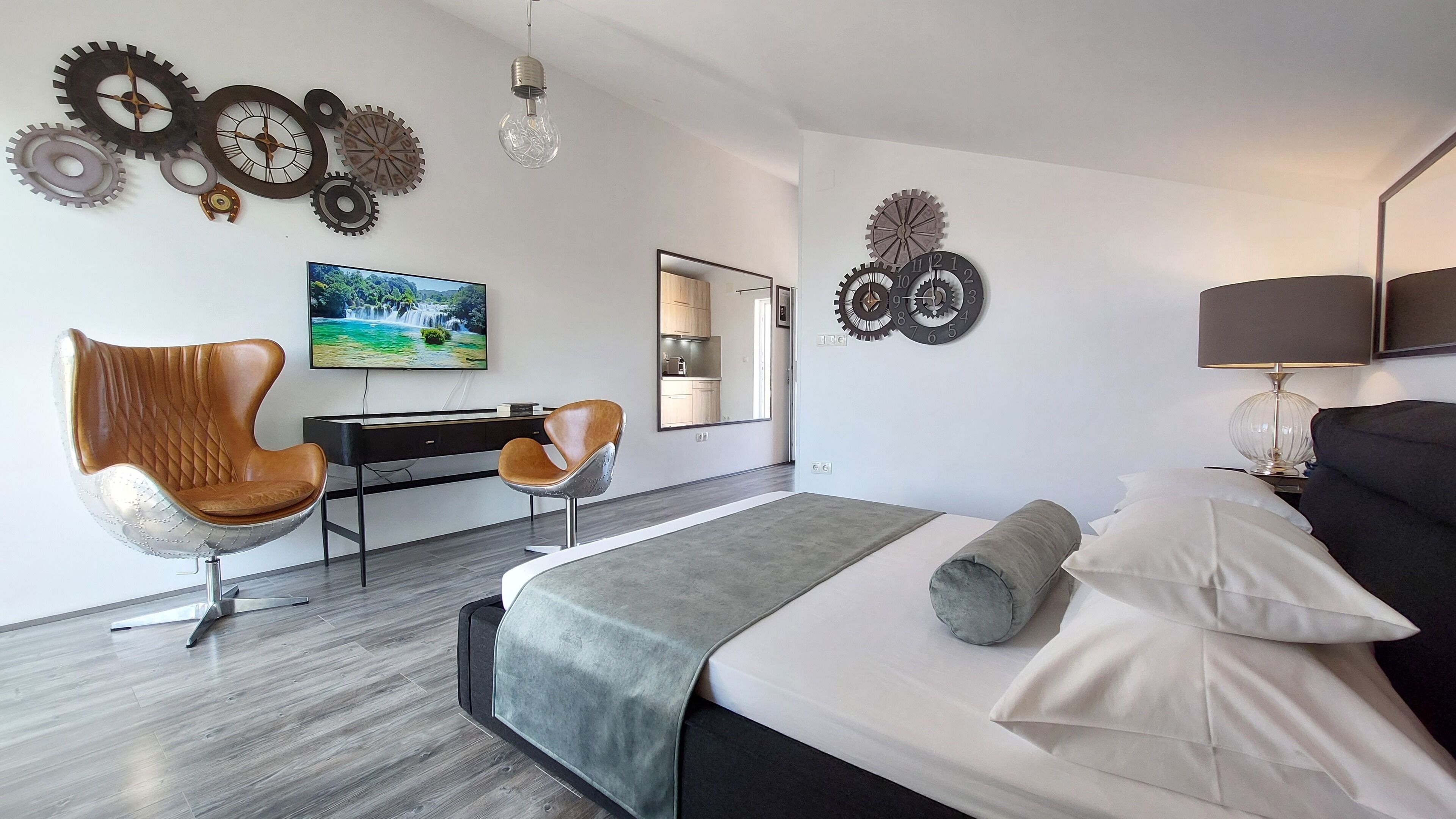 Una habitación de hotel moderna con cama, escritorio, dos sillas, televisor y relojes decorativos en la pared.