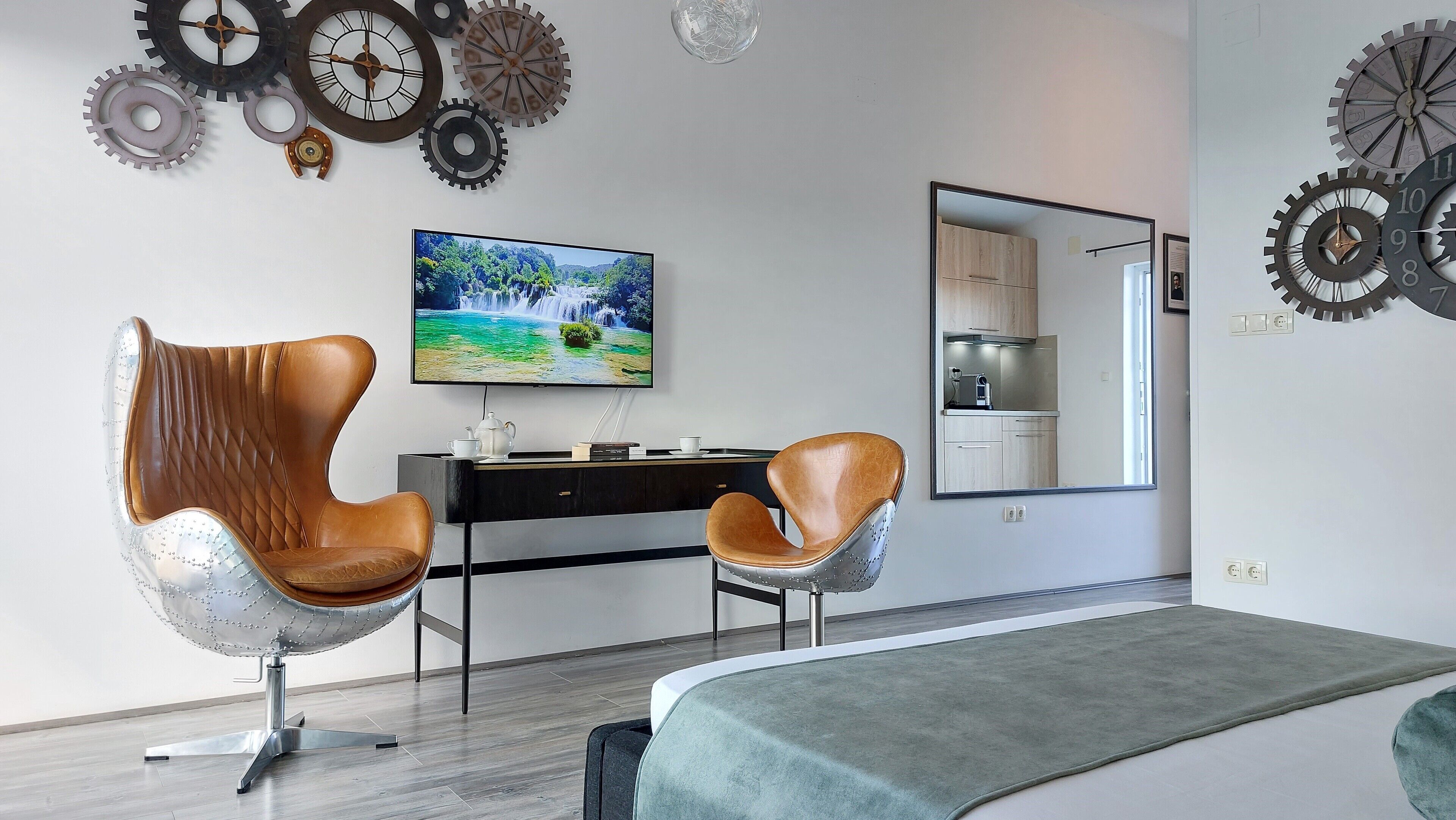 A modern bedroom with a bed, two leather chairs, a desk with a TV, and a kitchen area visible through a mirror.