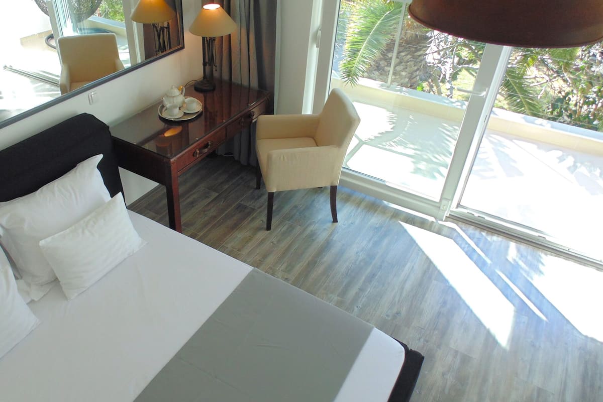 Habitación de hotel con una cama grande, un escritorio, una silla y un ventanal con vistas a palmeras.