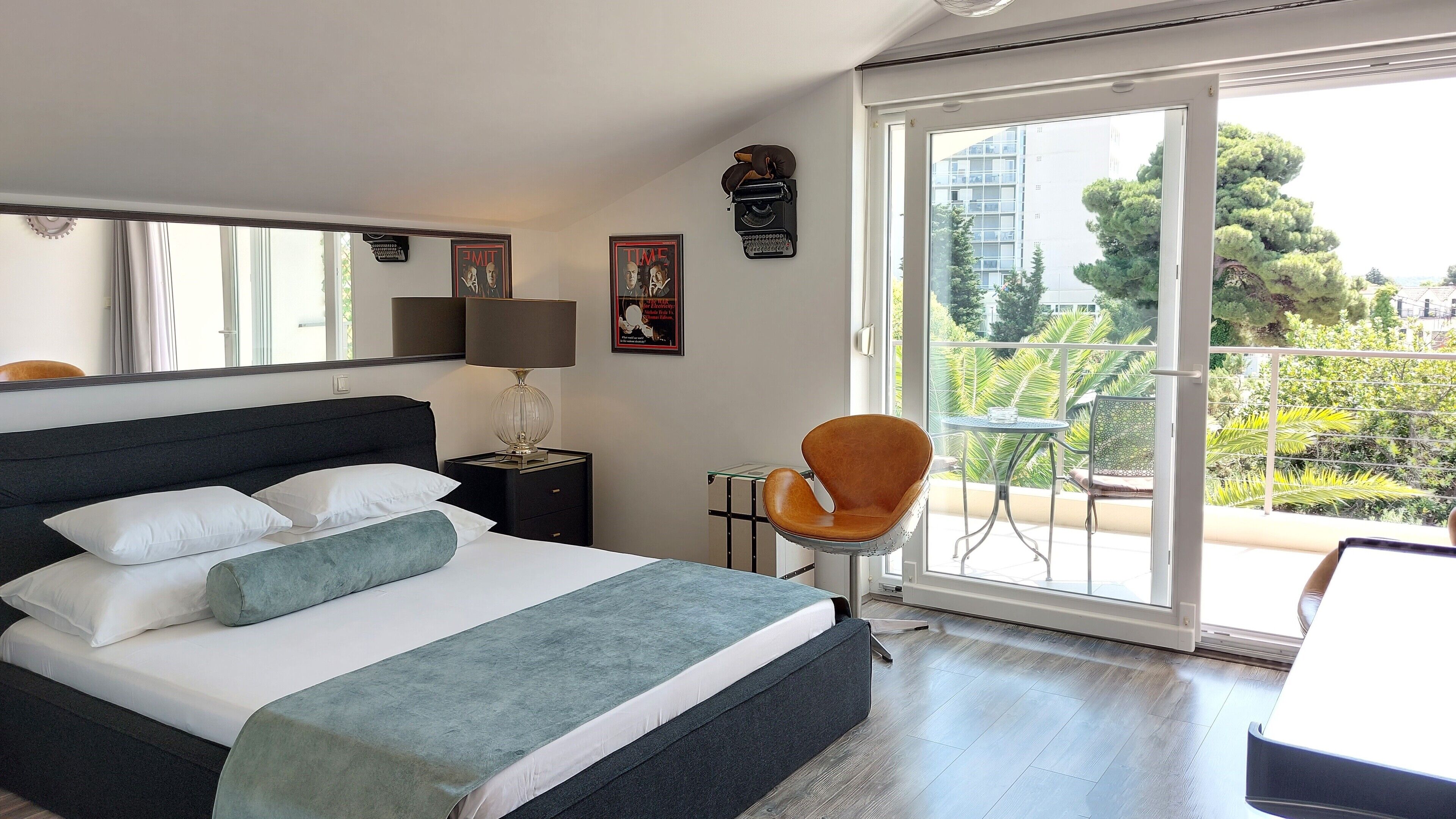 A hotel room with a large bed, a chair, a table, a balcony with a view of the city, and a large window.