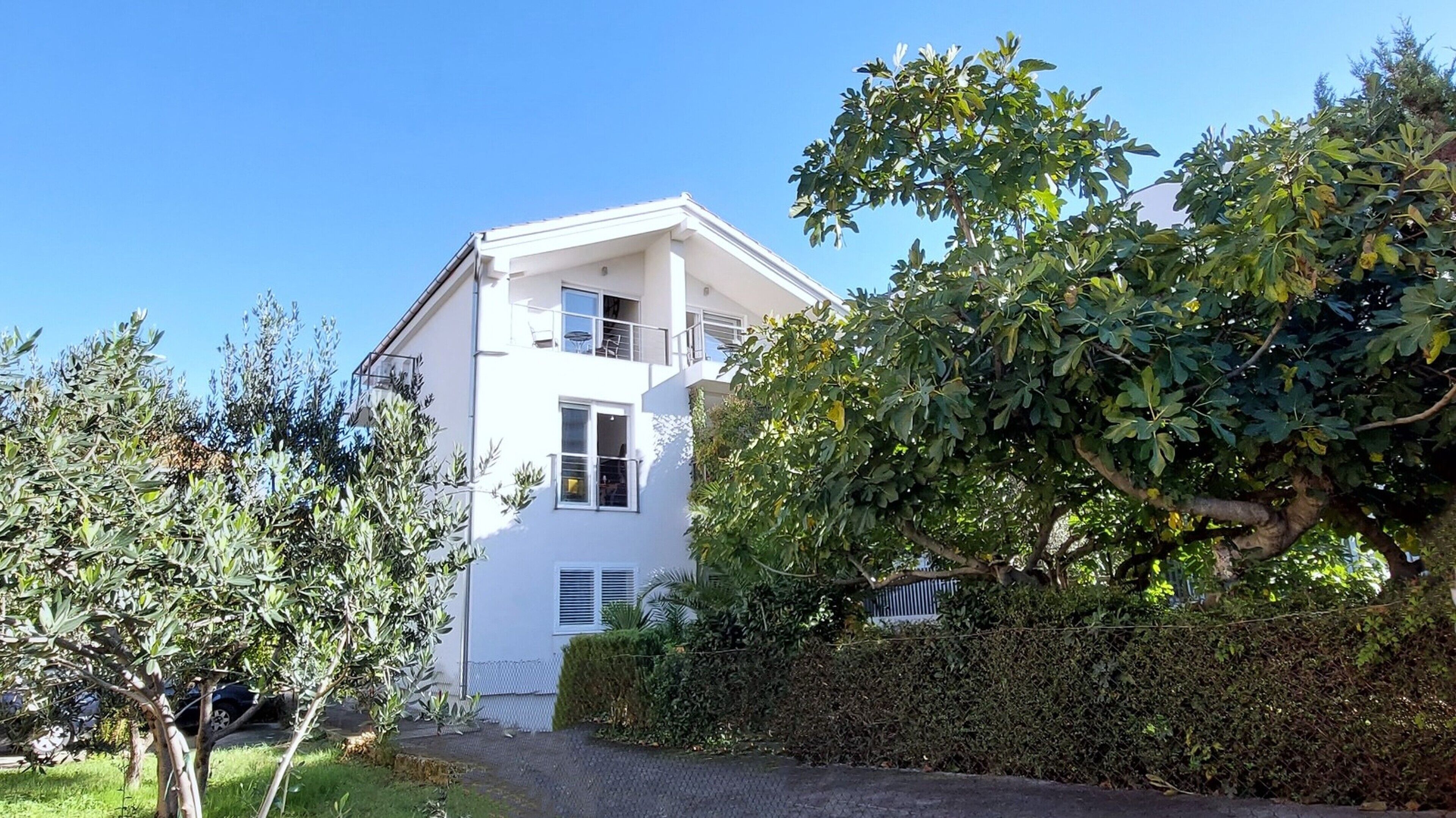 A two-story white building with a balcony, surrounded by greenery and a hedge.
