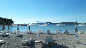 Una playa de guijarros con sillones blancos y sombrillas, un mar tranquilo y colinas al fondo.