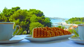 A table with waffles and two cups, overlooking a coastal view with trees and buildings. - Refresh Boutique Apartments (Vodice)