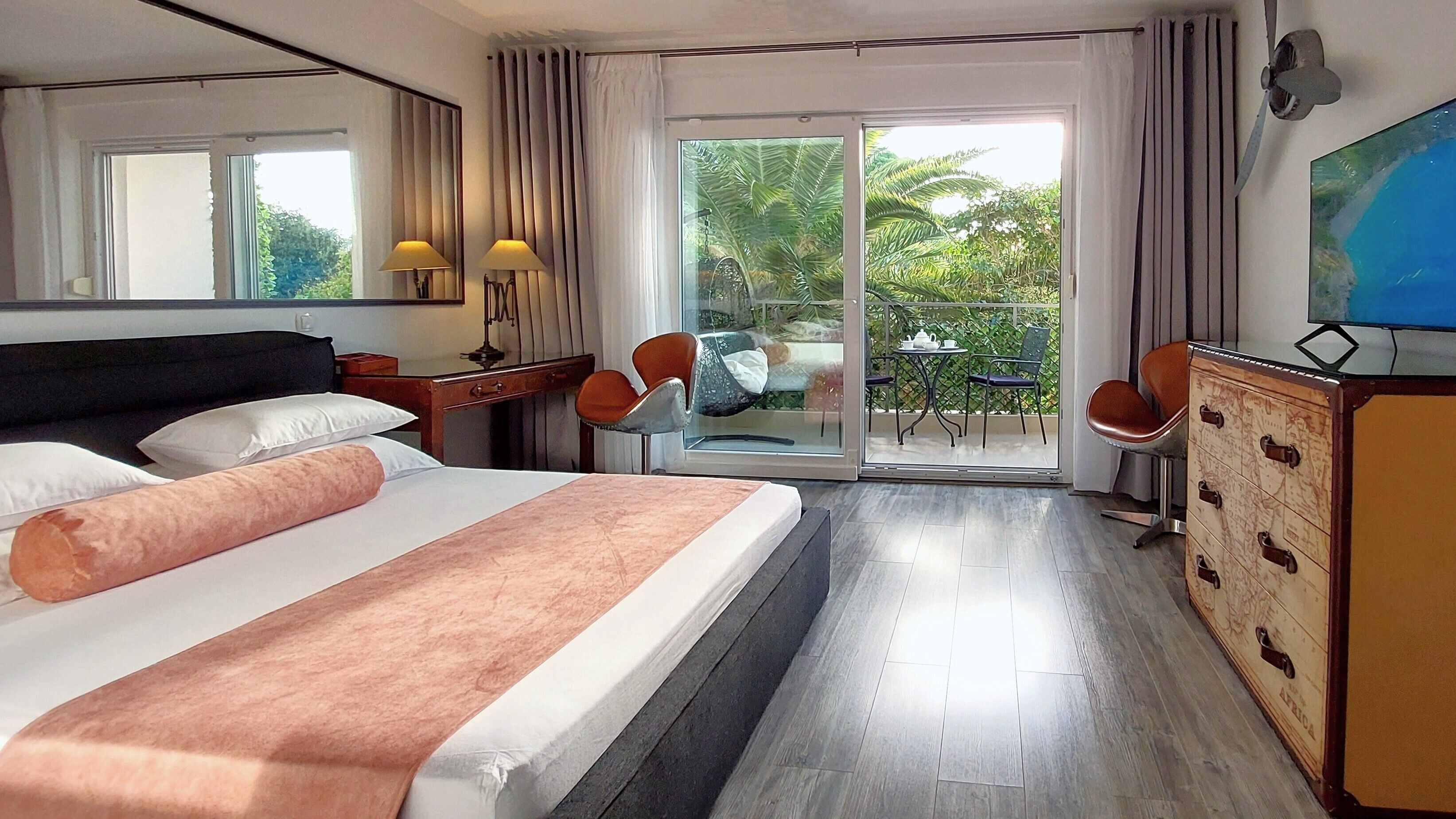 A hotel room with a bed, a desk, a chair, a dresser, and a sliding glass door leading to a balcony with outdoor furniture.