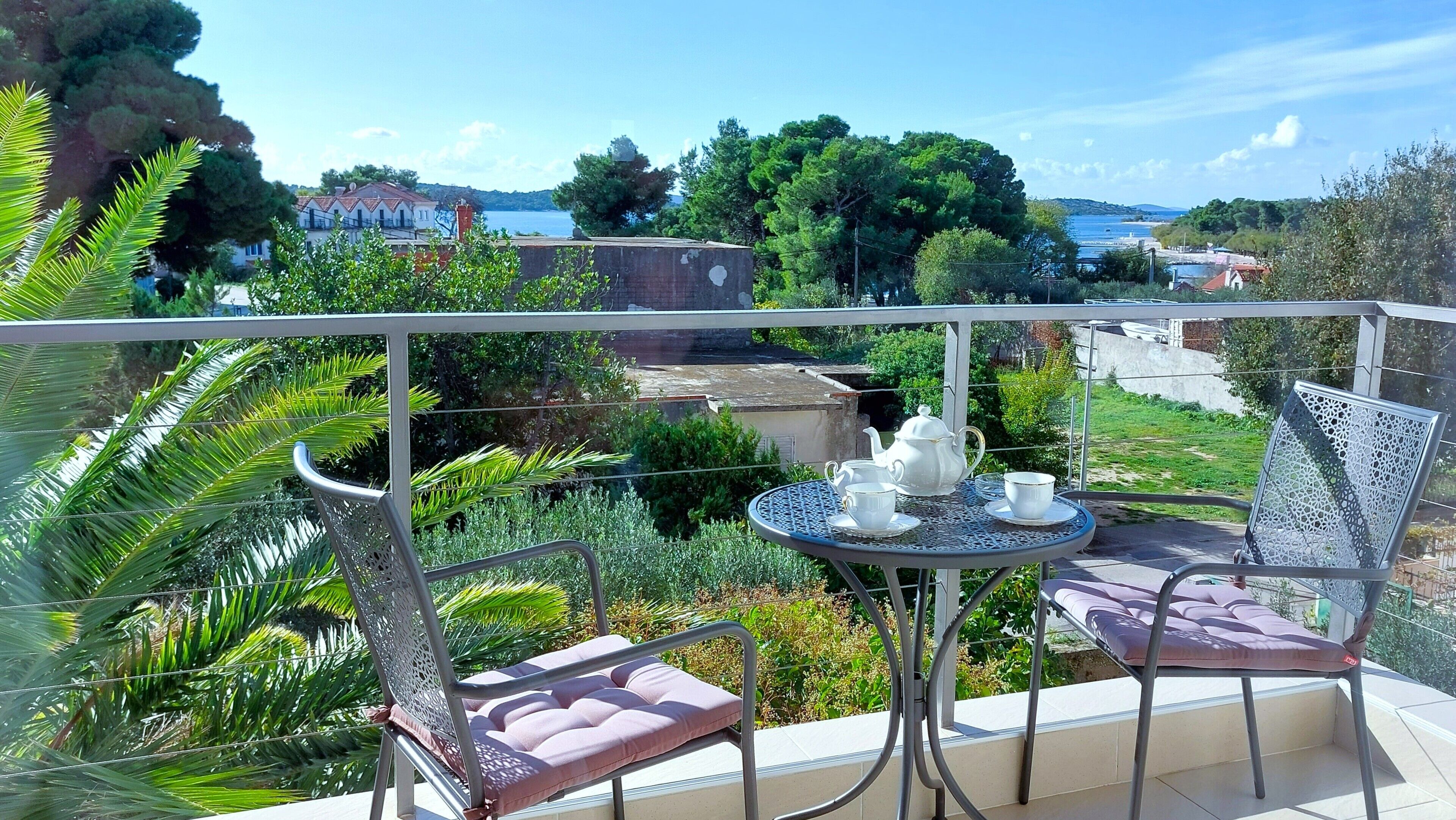 A balcony with a table set for tea, surrounded by lush greenery and a scenic view of a lake and buildings.