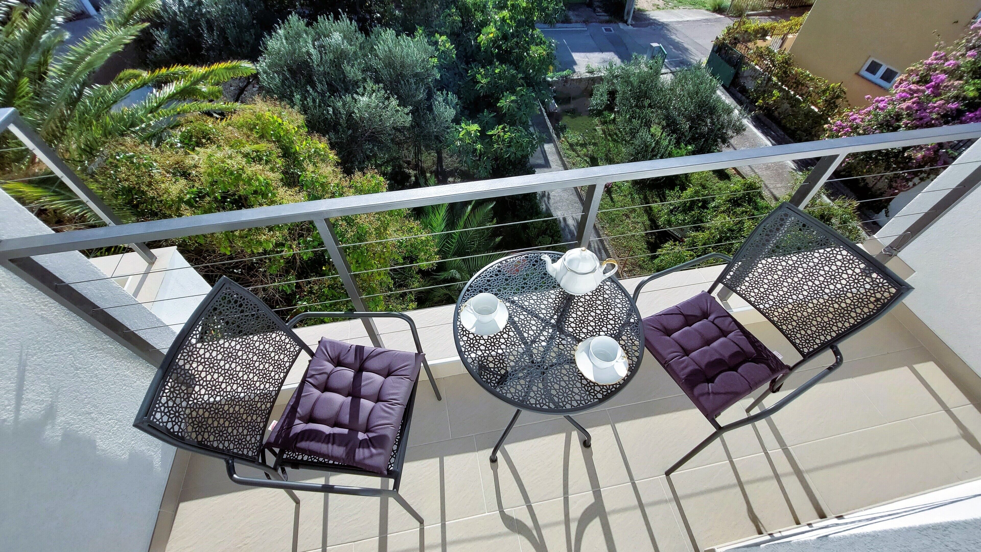 Un balcón con dos sillones acolchados y una pequeña mesa redonda, con vistas a un jardín exuberante y edificios colindantes.