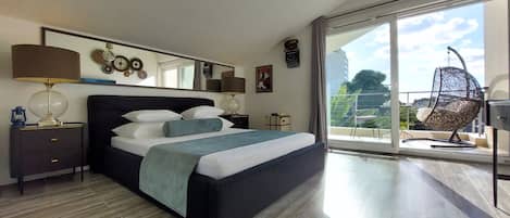 A modern bedroom with a large bed, a nightstand, a lamp, and a view of the outdoors through a sliding glass door.
