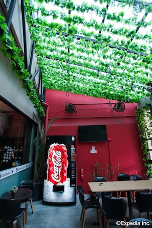 A restaurant with a red wall, a Coca-Cola vending machine, and green plants hanging from the ceiling. - K2 Guesthouse Central (Singapore)