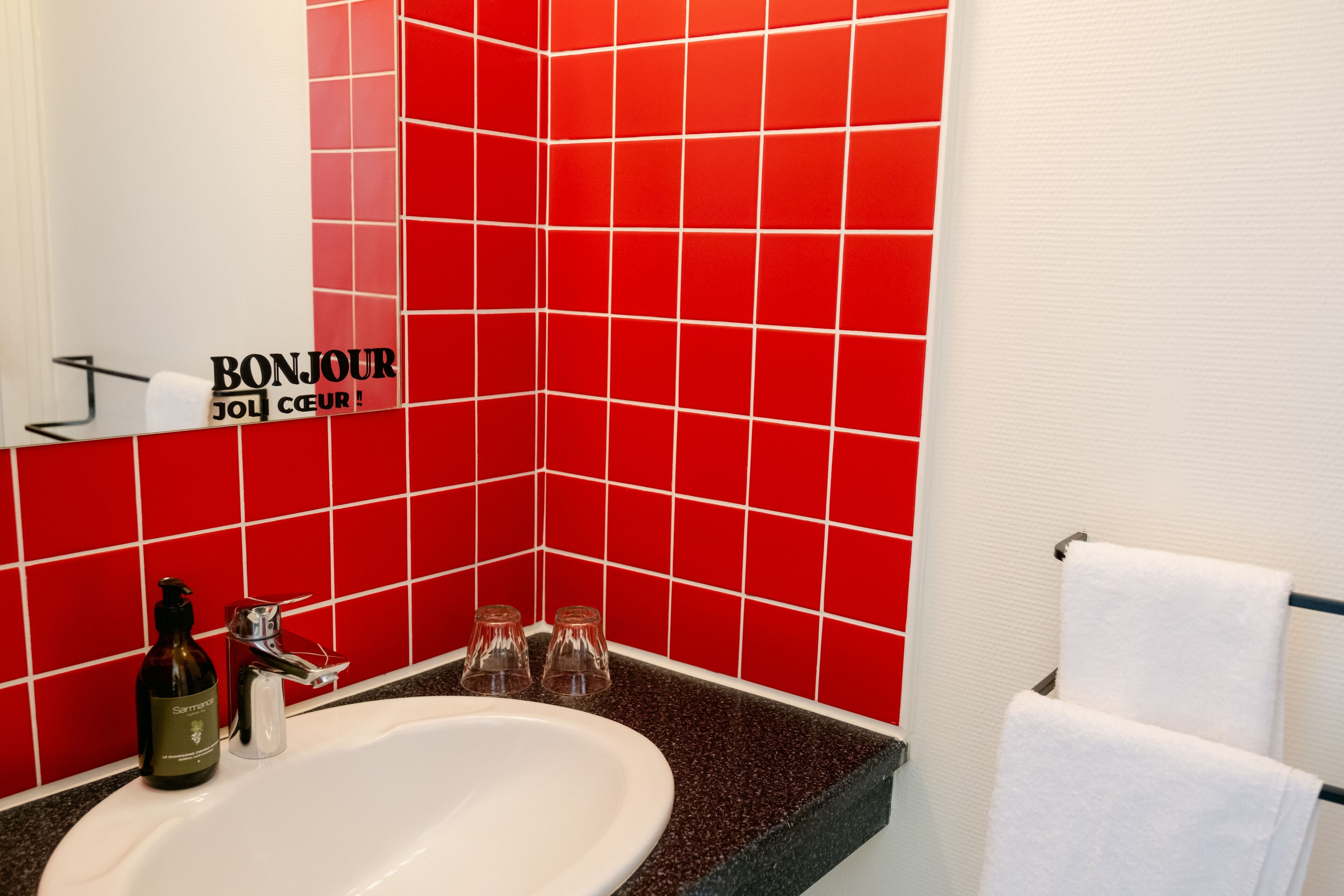 Un baño con pared de azulejos rojos, un lavamanos blanco, una botella de champú y un perchero para toallas.