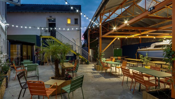 An outdoor patio area with tables and chairs, a staircase, and a boat visible in the background. - Le Lieu Dit (Nantes)