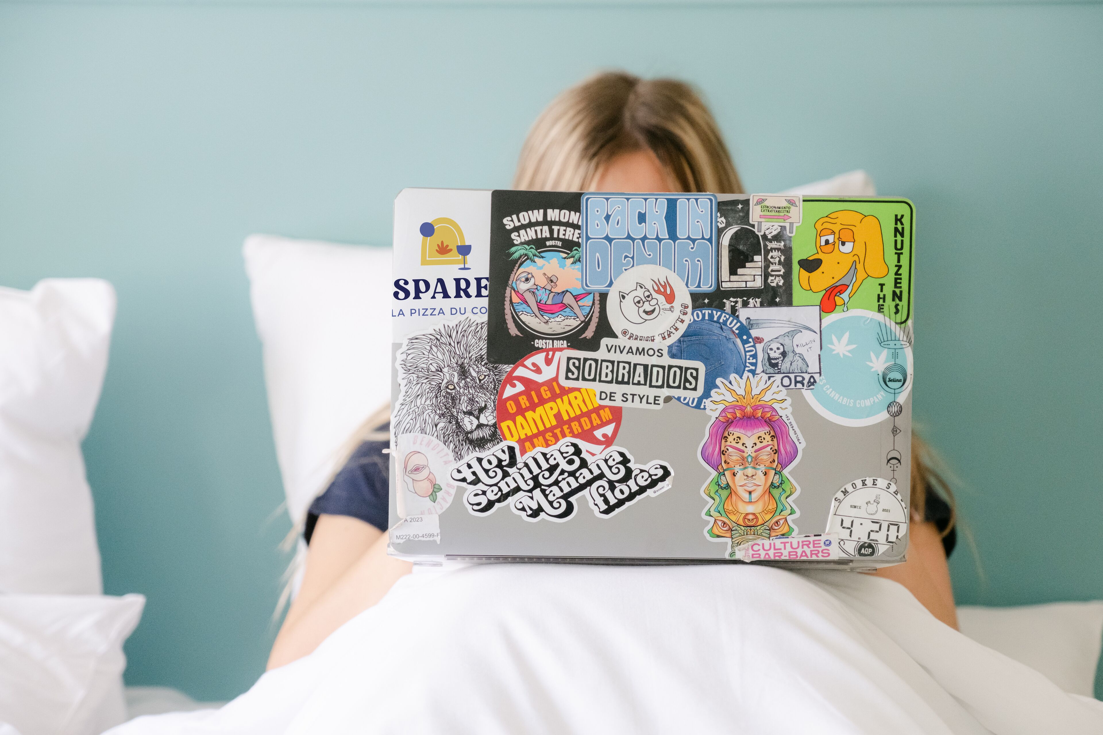 A person lying on a bed with a laptop covered in various stickers.