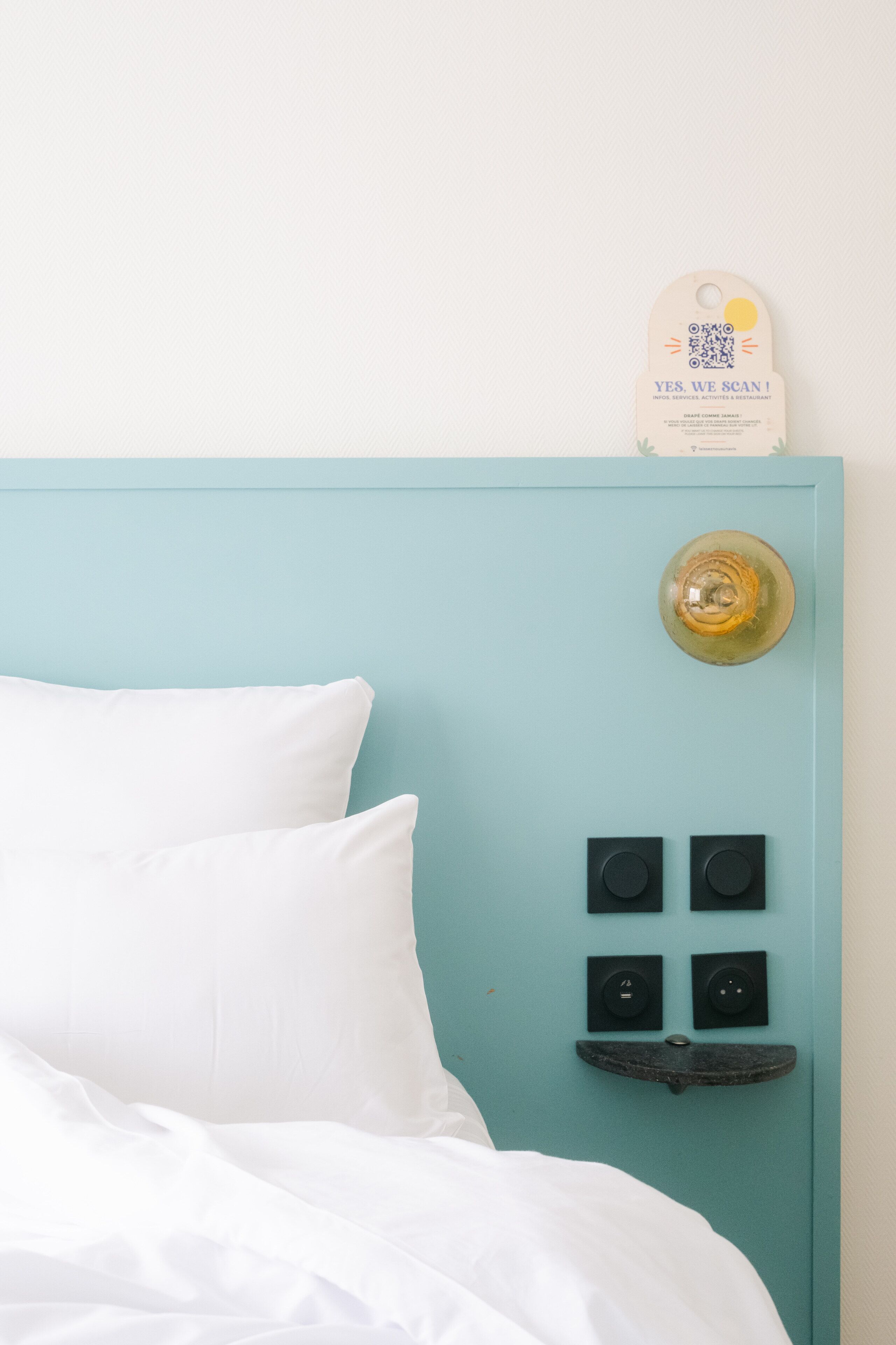 Una cama con sábanas blancas, un cabecero color turquesa con un código QR y un espejo redondo pequeño.