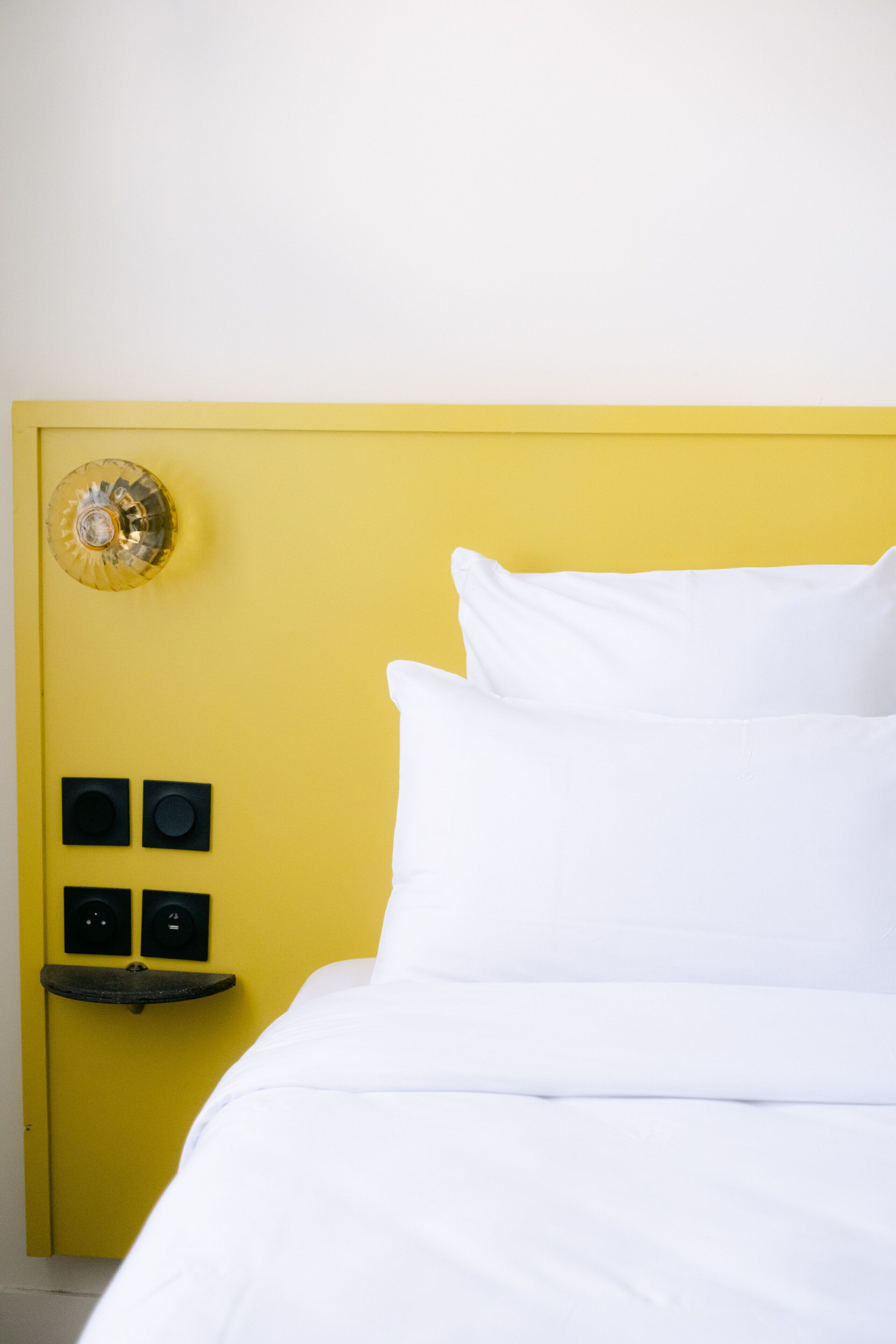 Ein Bett mit weißer Bettwäsche in einem Zimmer, das ein gelbes Kopfende und einen kleinen Nachttisch mit einer Lampe hat.