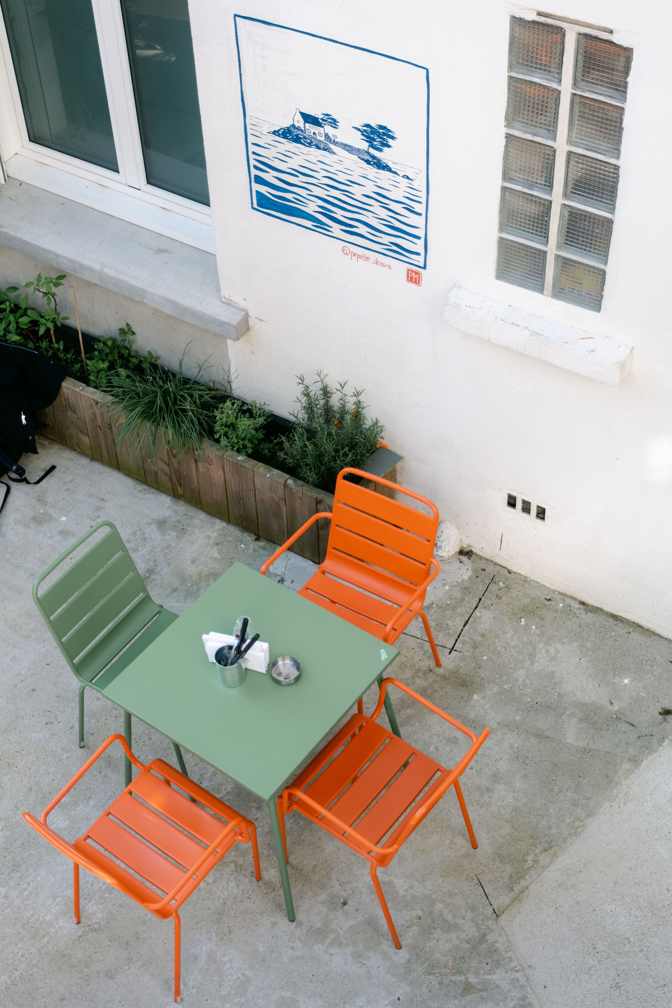 An outdoor patio with a green table and orange chairs, a poster on the wall, and a wooden planter with plants.
