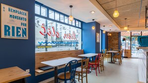 A restaurant interior with wooden tables and chairs, a large window with a view of the street, and a neon sign on the wall. - Le Lieu Dit (Nantes)