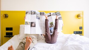 Two people lying on a bed with a yellow headboard, reading a magazine. - Le Lieu Dit (Nantes)