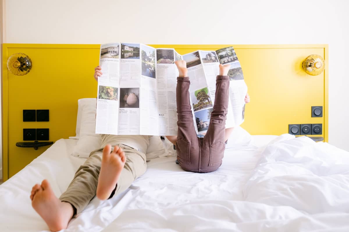 Dos personas recostadas en una cama con cabecera amarilla, leyendo una revista.
