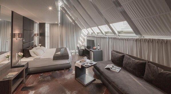 A modern hotel room with a large bed, a sofa, a desk, and a dining table. There is a TV, a laptop, and a window with curtains.