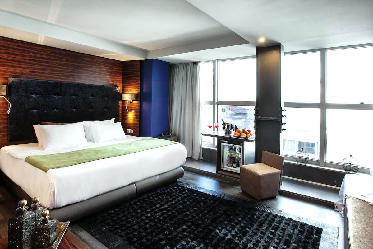 A modern hotel room with a large bed, a small table with fruit, a chair, and a view of the city through large windows.