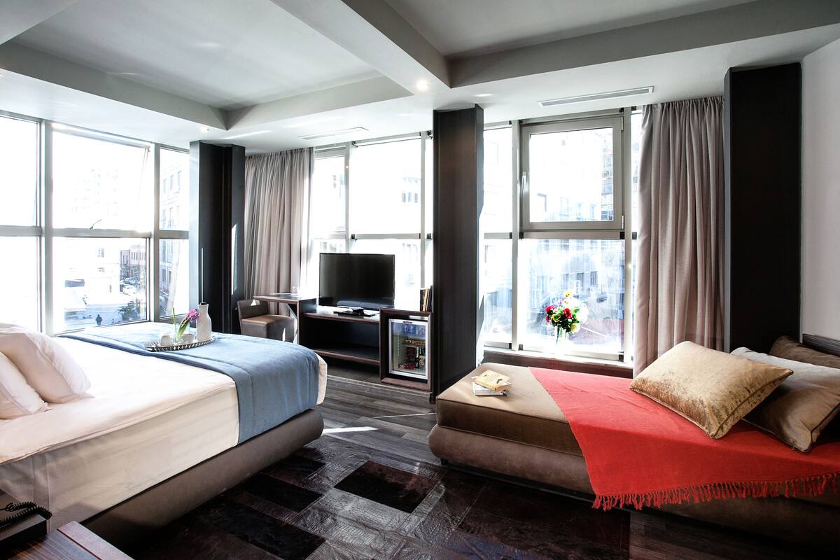 A hotel room with two beds, a TV, a desk, and large windows with curtains.