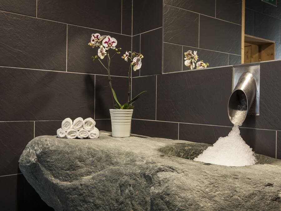 Un baño con baldosas oscuras, un lavabo de piedra, una orquídea en maceta y toallas enrolladas con cuidado.