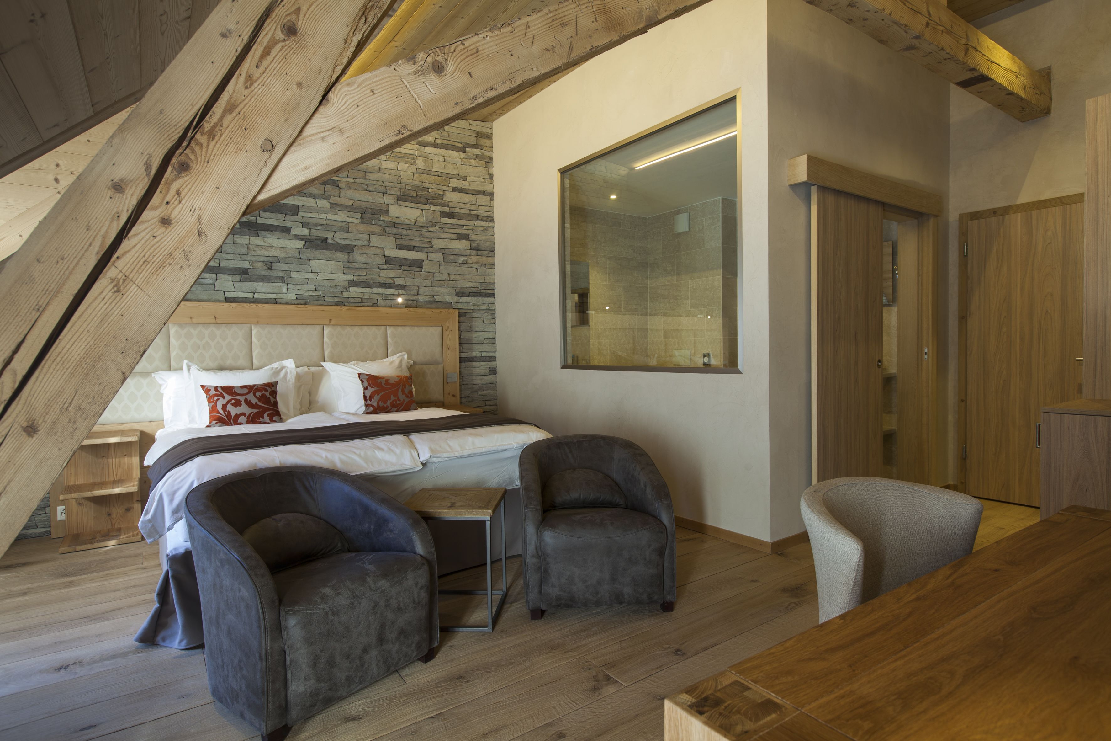 A bedroom with a sloped ceiling, a large bed, two armchairs, a wooden table, and a wardrobe.