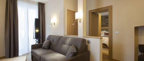 A modern living room with a grey sofa, wooden floor, and a mirror reflecting a bedroom.