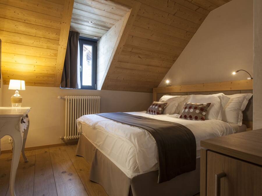 Un dormitorio con techo inclinado, una cama con ropa blanca y una manta marrón, una mesita de noche con lámpara y una ventana con cortinas.