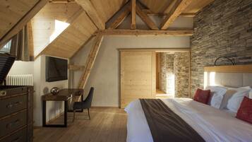 A bedroom with a sloped ceiling, wooden beams, a stone wall, a bed with white and red pillows, a desk with a chair, and a dresser.