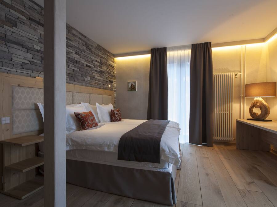 A modern hotel room with a stone accent wall, a large bed with multiple pillows, a bedside table with a lamp, and a wooden bench.