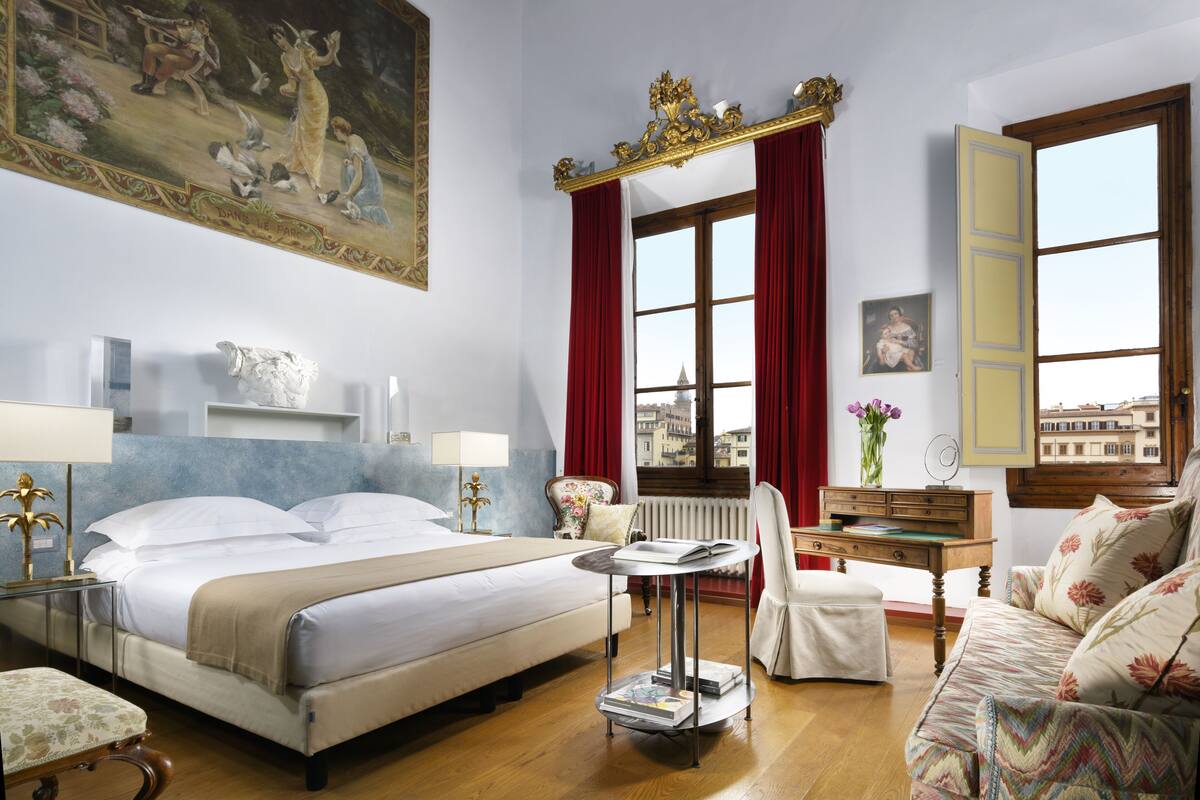 A hotel room with a bed, a desk, a chair, a sofa, a window with curtains, and a painting on the wall.