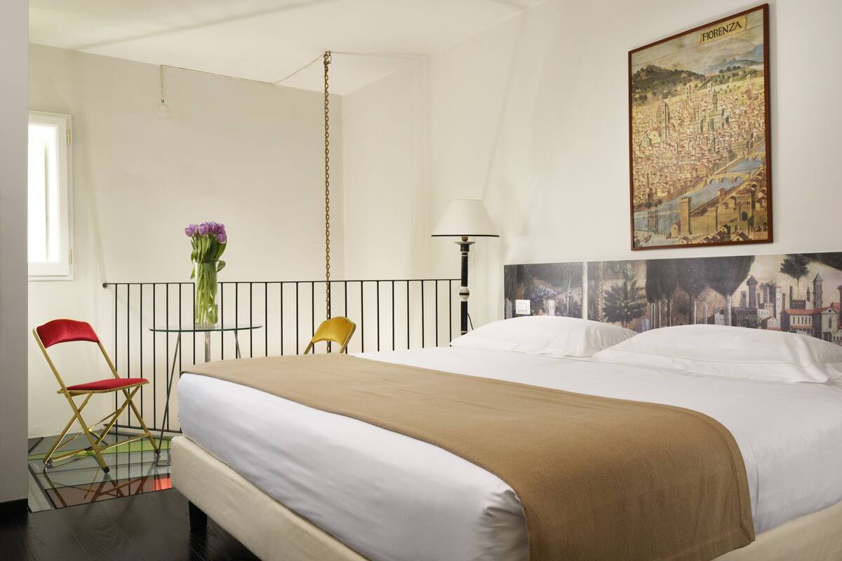 A hotel room with a bed, a red chair, a yellow chair, a table with a vase of flowers, a lamp, and a painting of Florence on the wall.