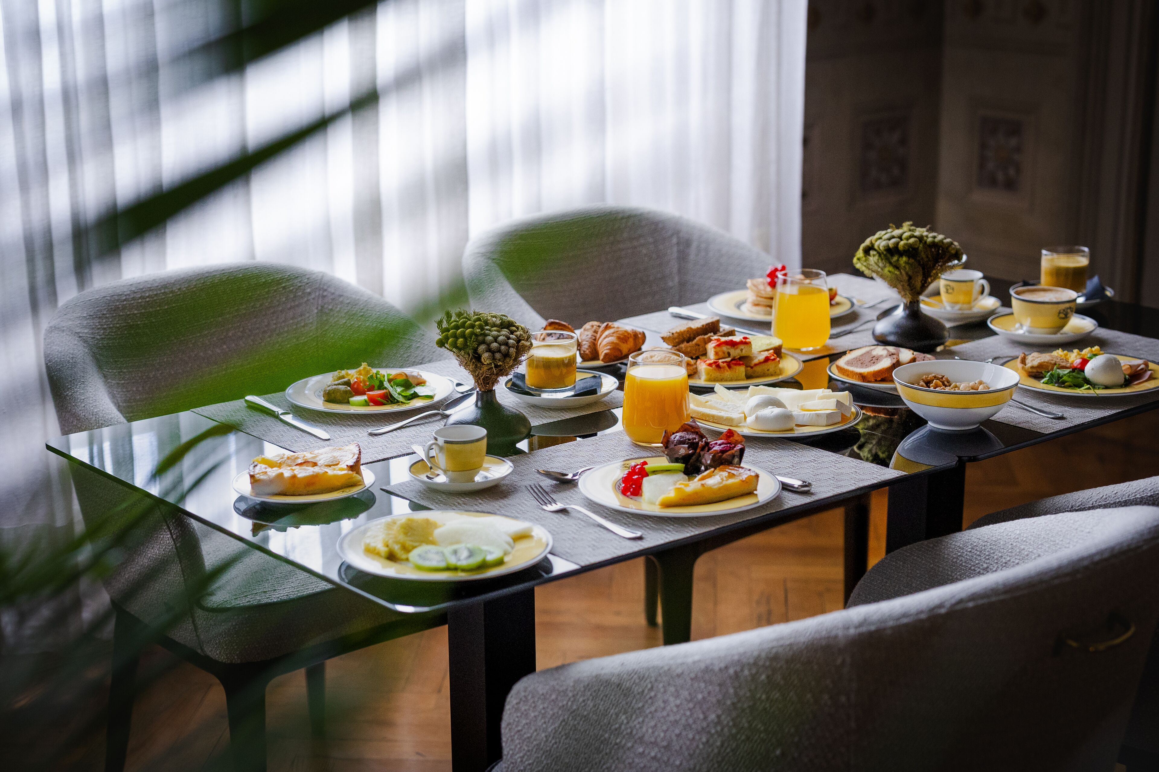A breakfast table set with various dishes, including pastries, fruits, and beverages, in a room with modern furniture and a view of the outdoors.