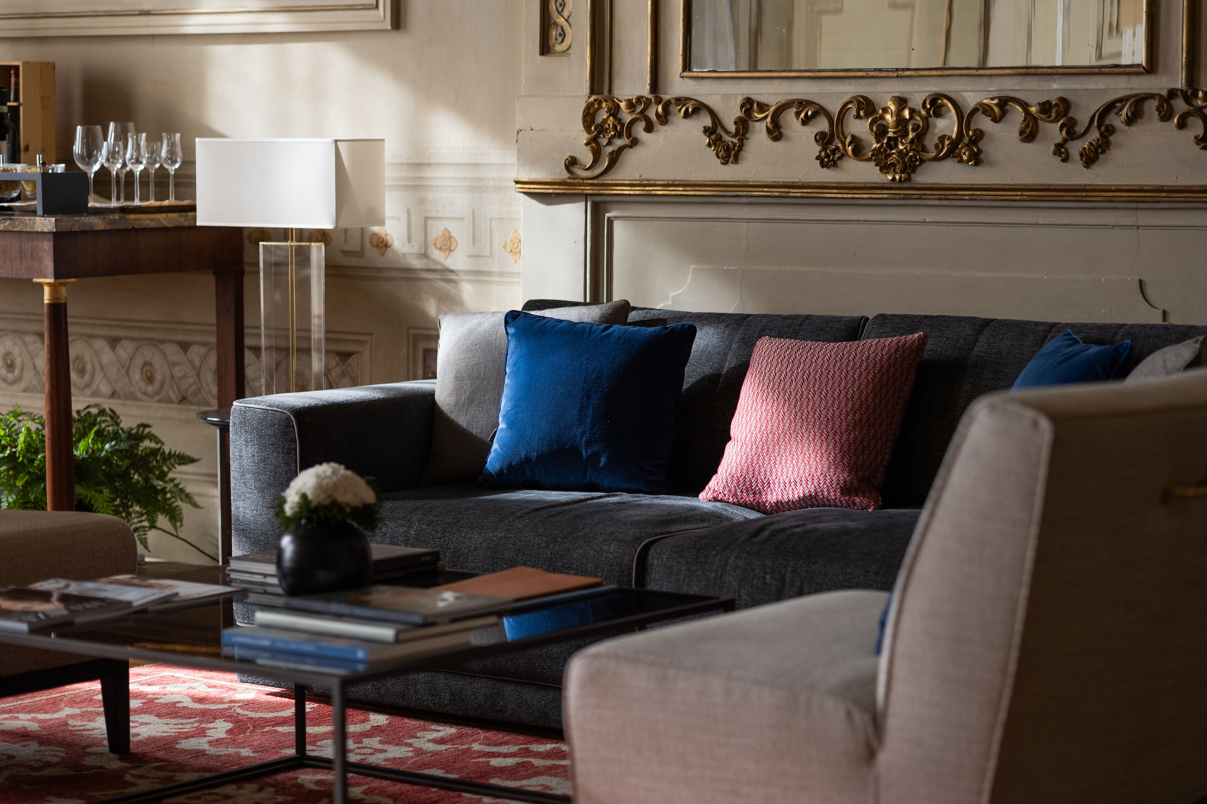 A modern living room with a grey sofa, blue and pink cushions, a coffee table with books and a vase, and a lamp on a side table.